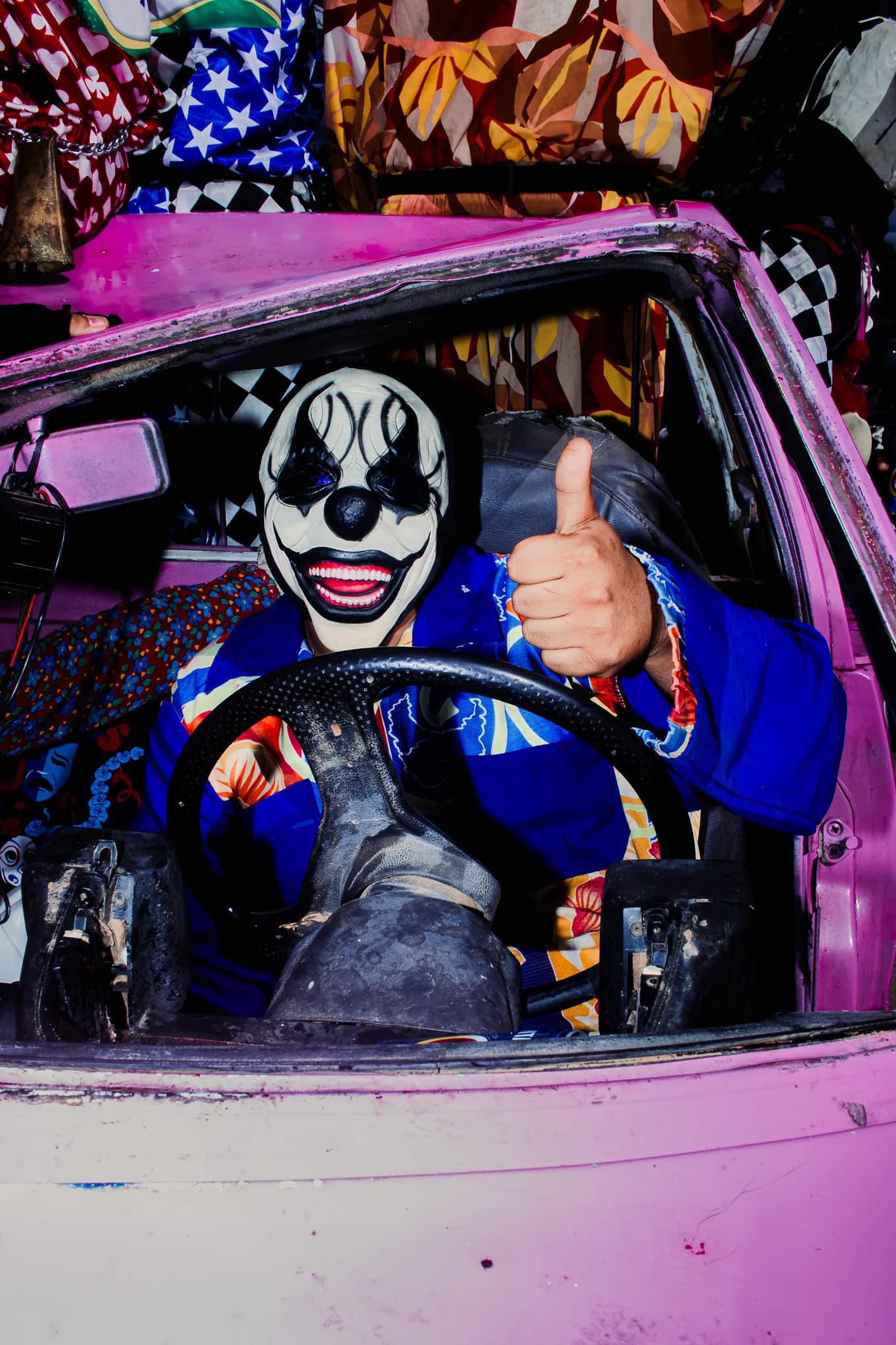 A masked clown at the wheel of a pink car, giving a thumbs up.