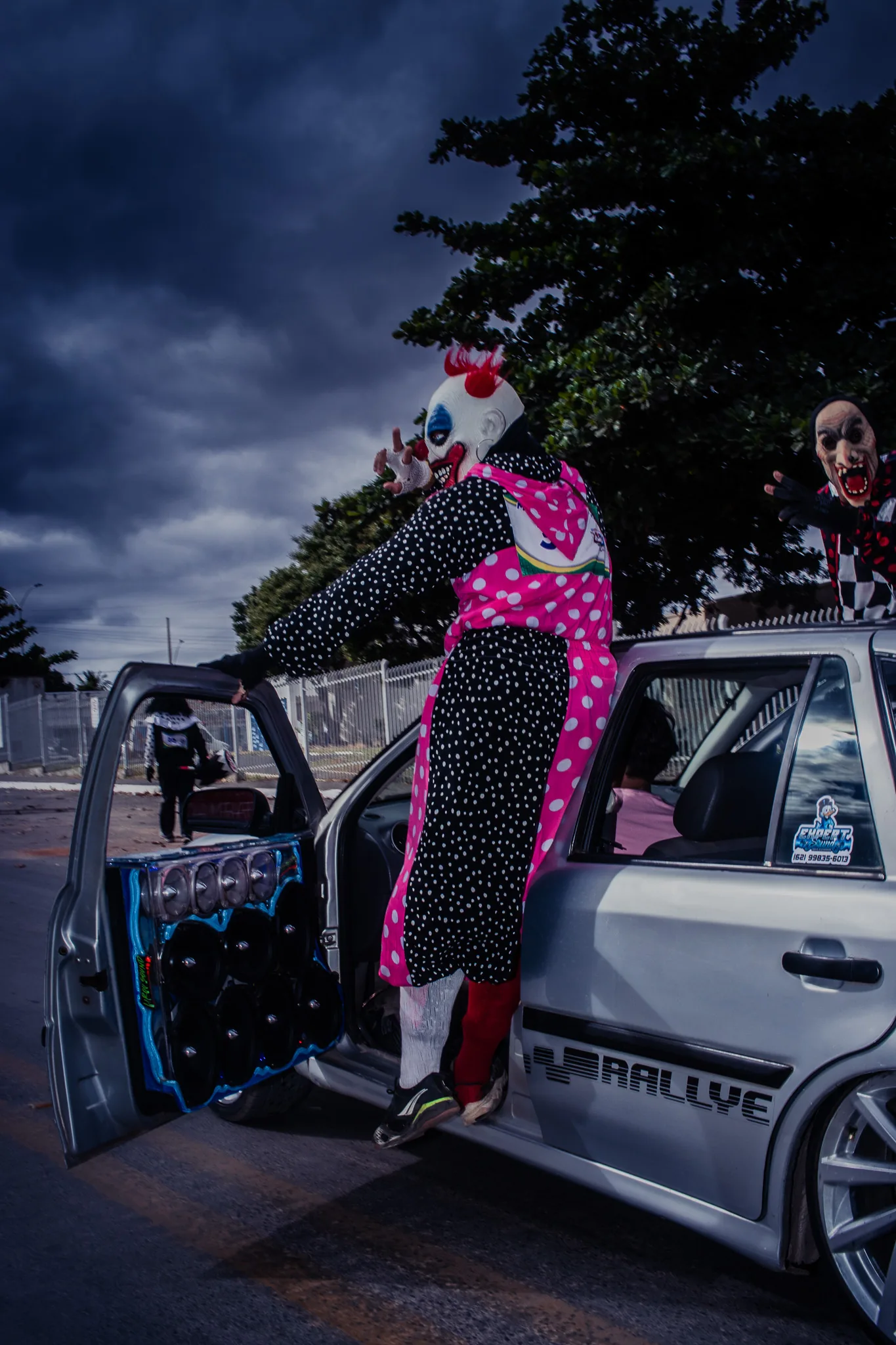 A clown in a polka dot costume standing on the door of a white car with the sound system open.