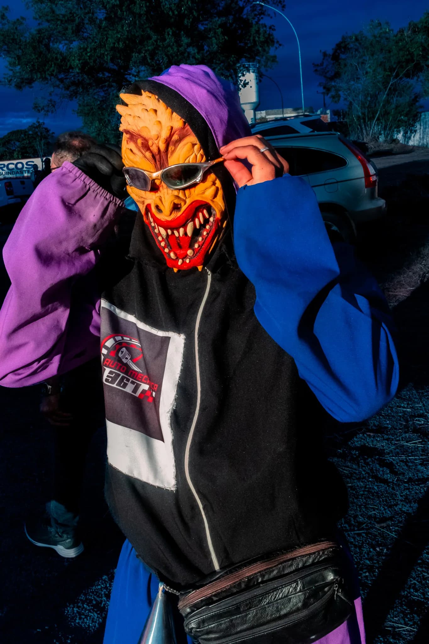 A young man pulls an orange devil mask over his face, leaning on a car.