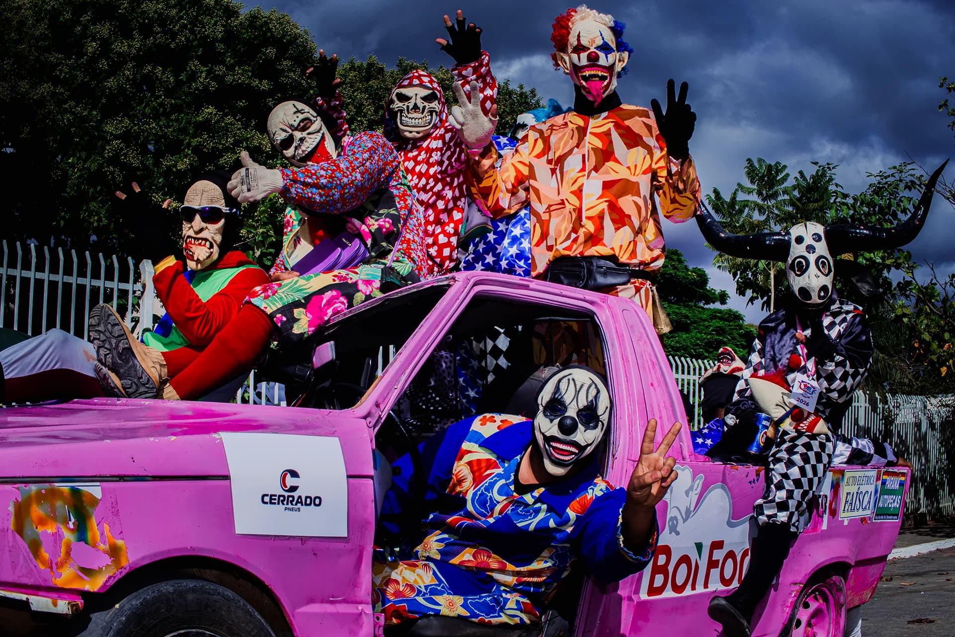 Foliões mascarados sobre um carro rosa na festa de Posse: máscaras de palhaço, caveira e boi, mãos erguidas, céu de tempestade atrás.