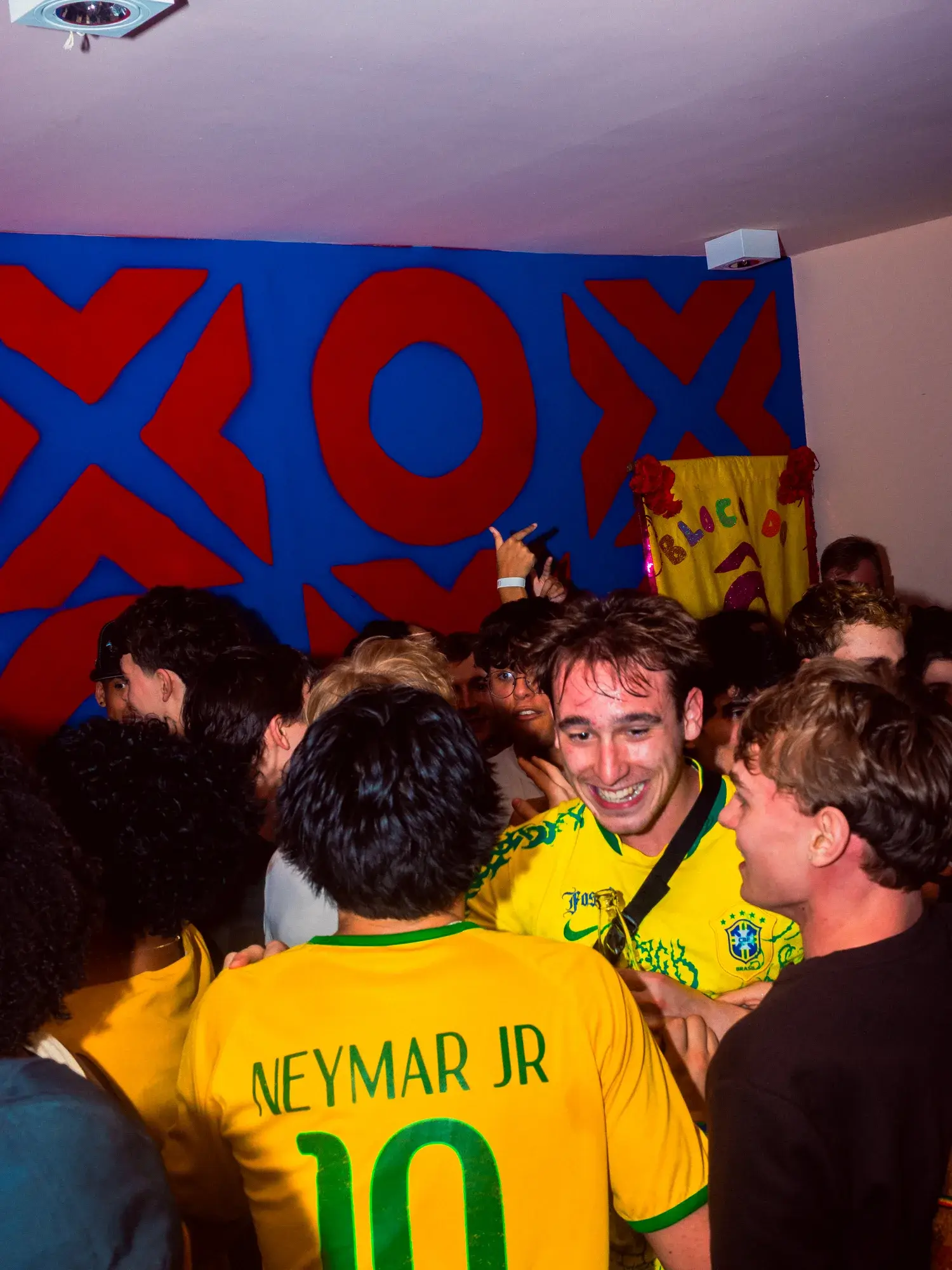 Guests crowded together watching a match in Brazil shirts, under the painted XOXO wall.