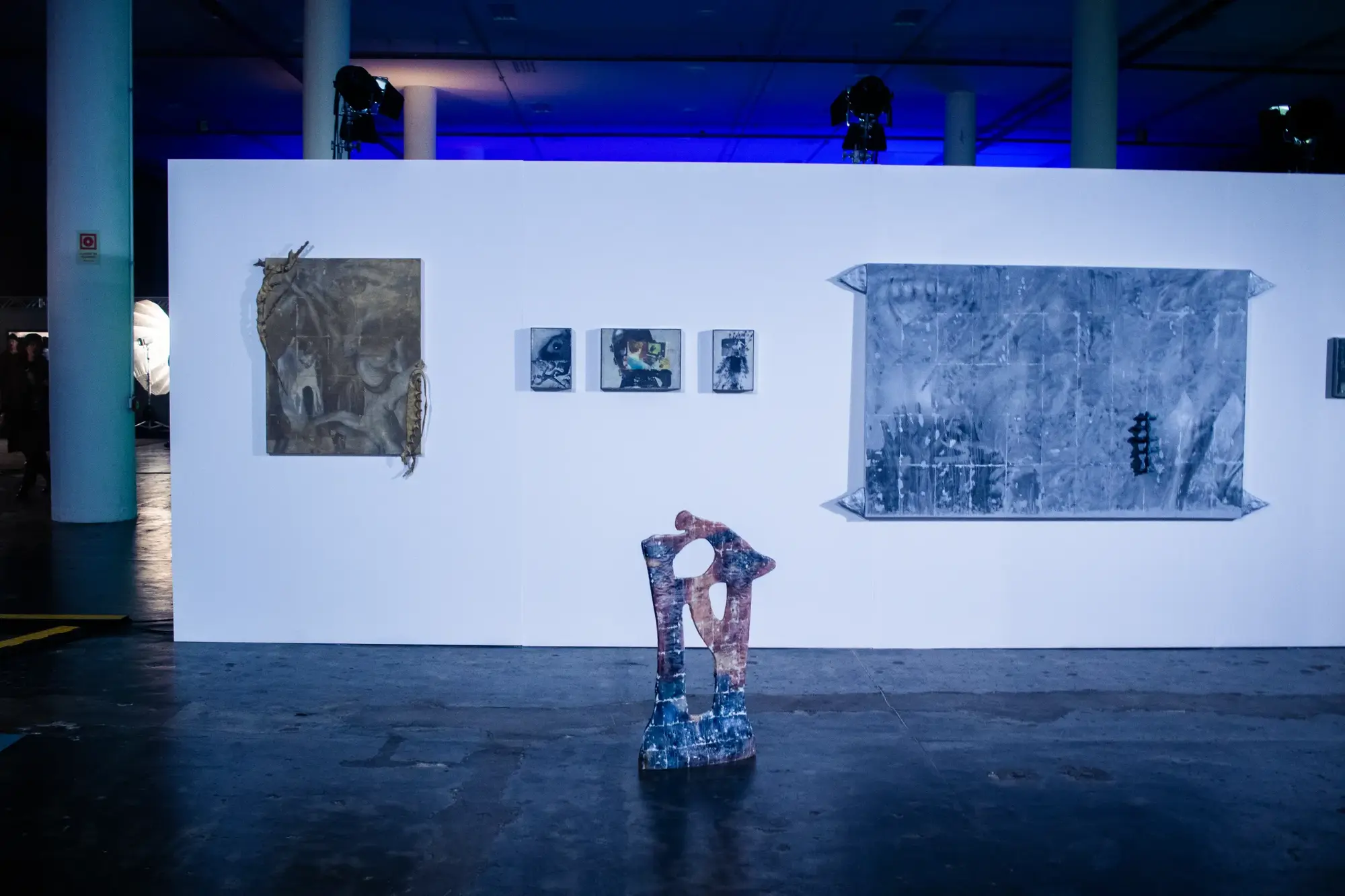 A gallery wall with paintings and a sculpture in a blue-lit exhibition hall.